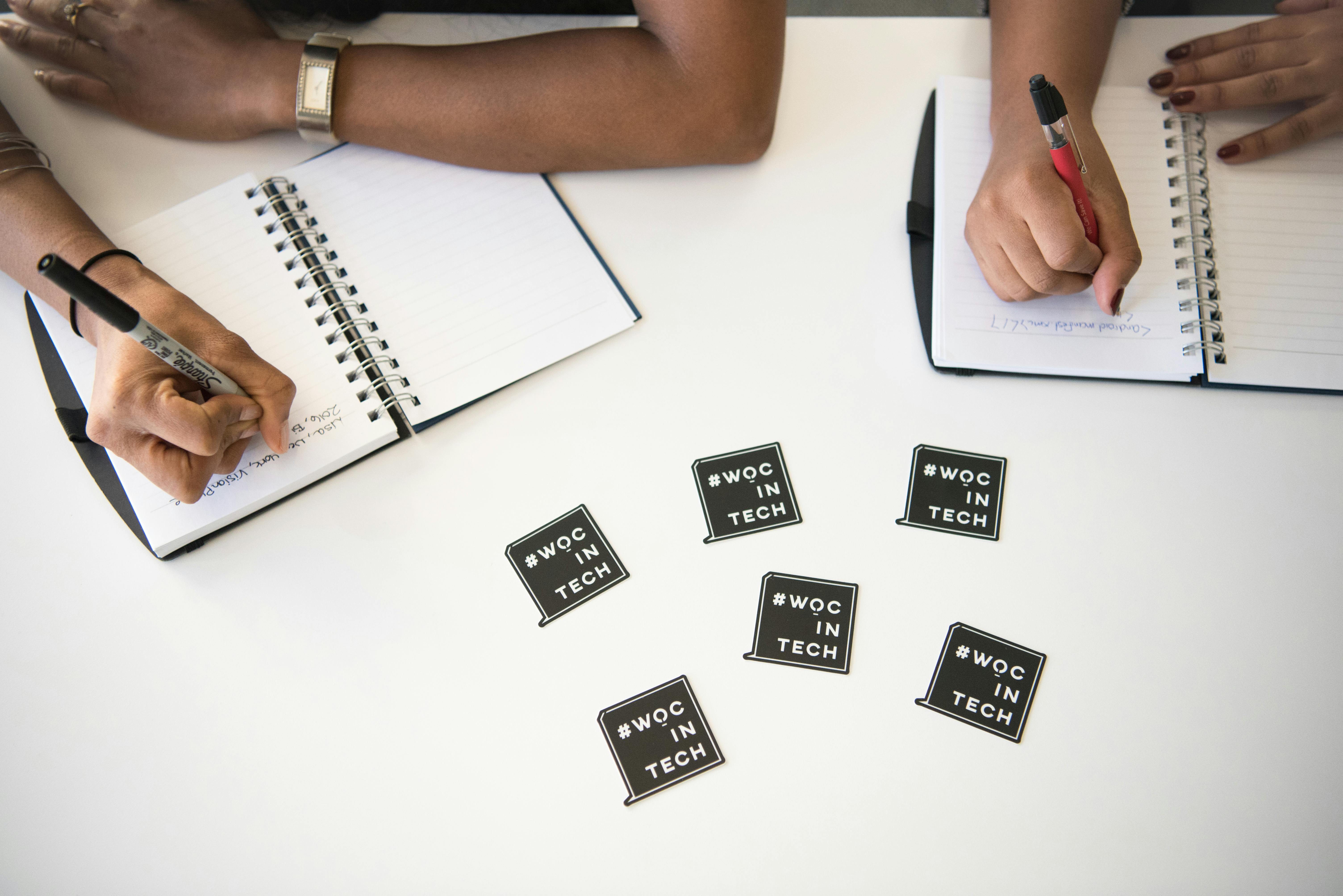 Diverse women collaborating in a tech meeting, writing notes in notebooks on a white desk.